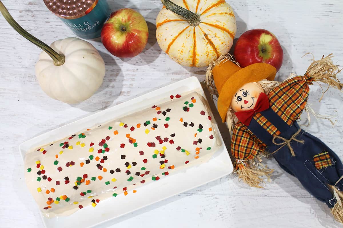 Overhead fall sprinkled roll cake on white platter with pumpkins, apples and scarecrow around it.