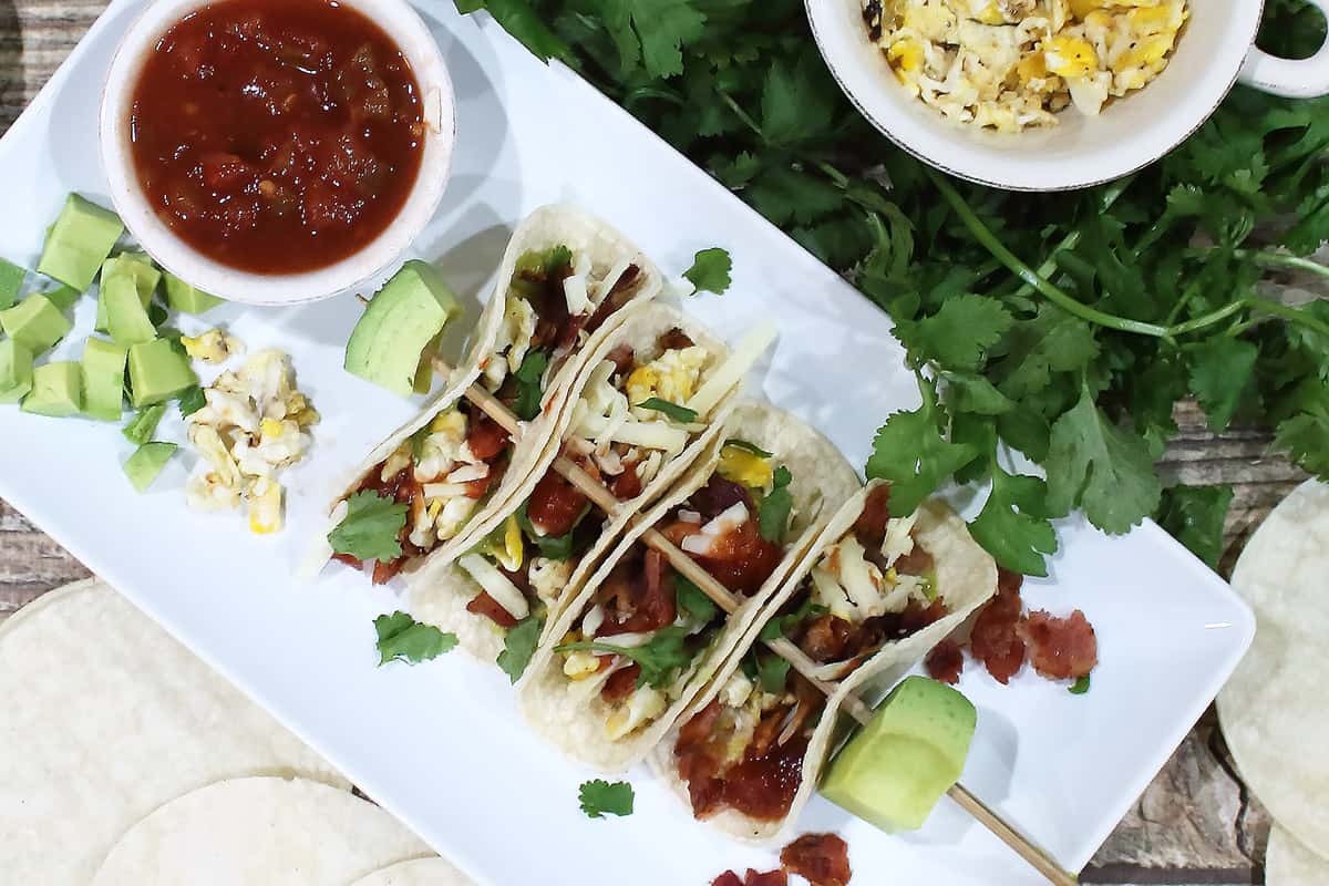 Overhead of skewered bacon and egg tacos on white platter with salsa and avocado.