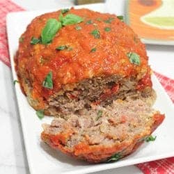 Meatloaf on serving plate with one slice and basil garnish.