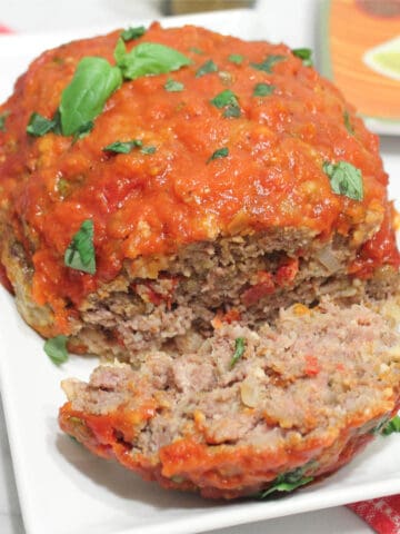 Meatloaf on serving plate with one slice and basil garnish.