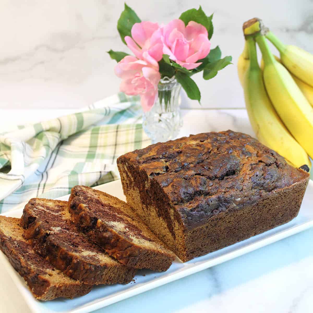 Banana bread on white platter with flowers and bunch of bananas.