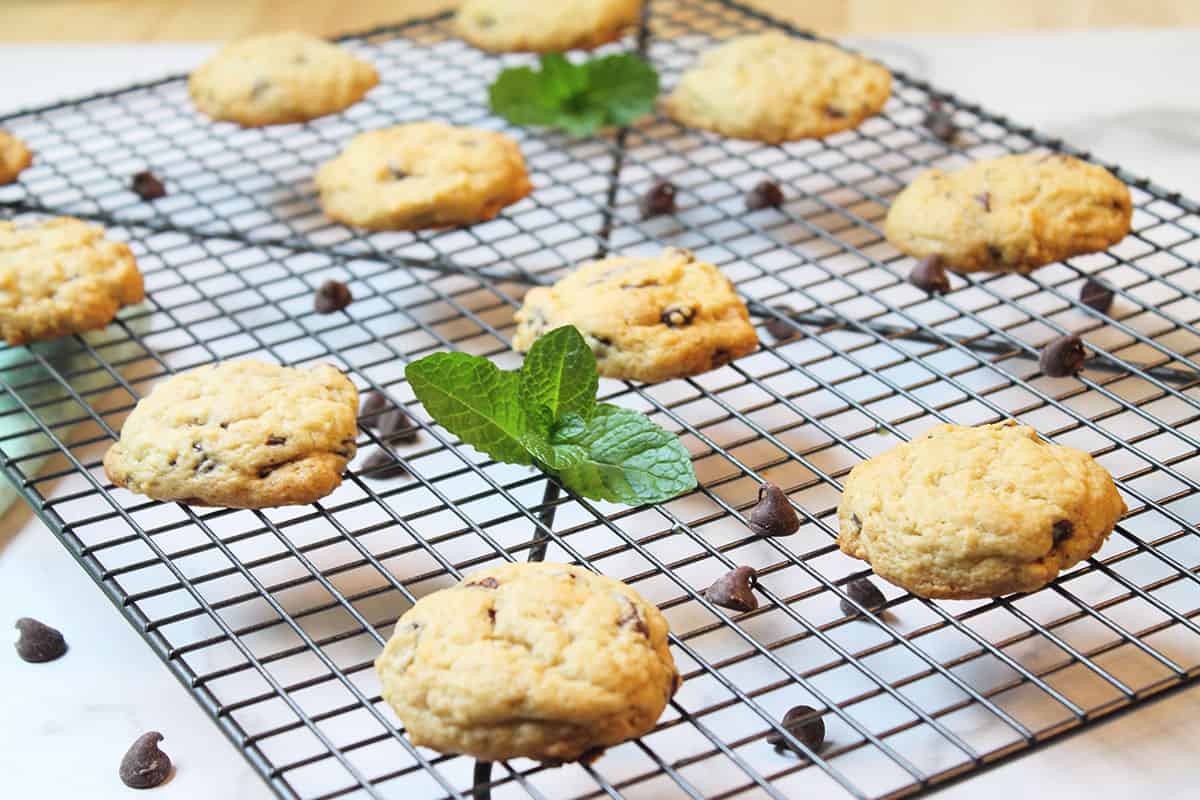 Mint chip cookies cooling on wire rack with mint sprigs and chocolate chips scattered around.