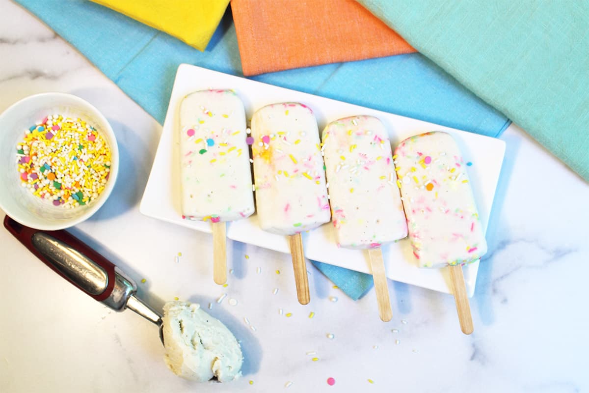 Overhead of cake pops on plate with colorful napkins in back and sprinkles on side.