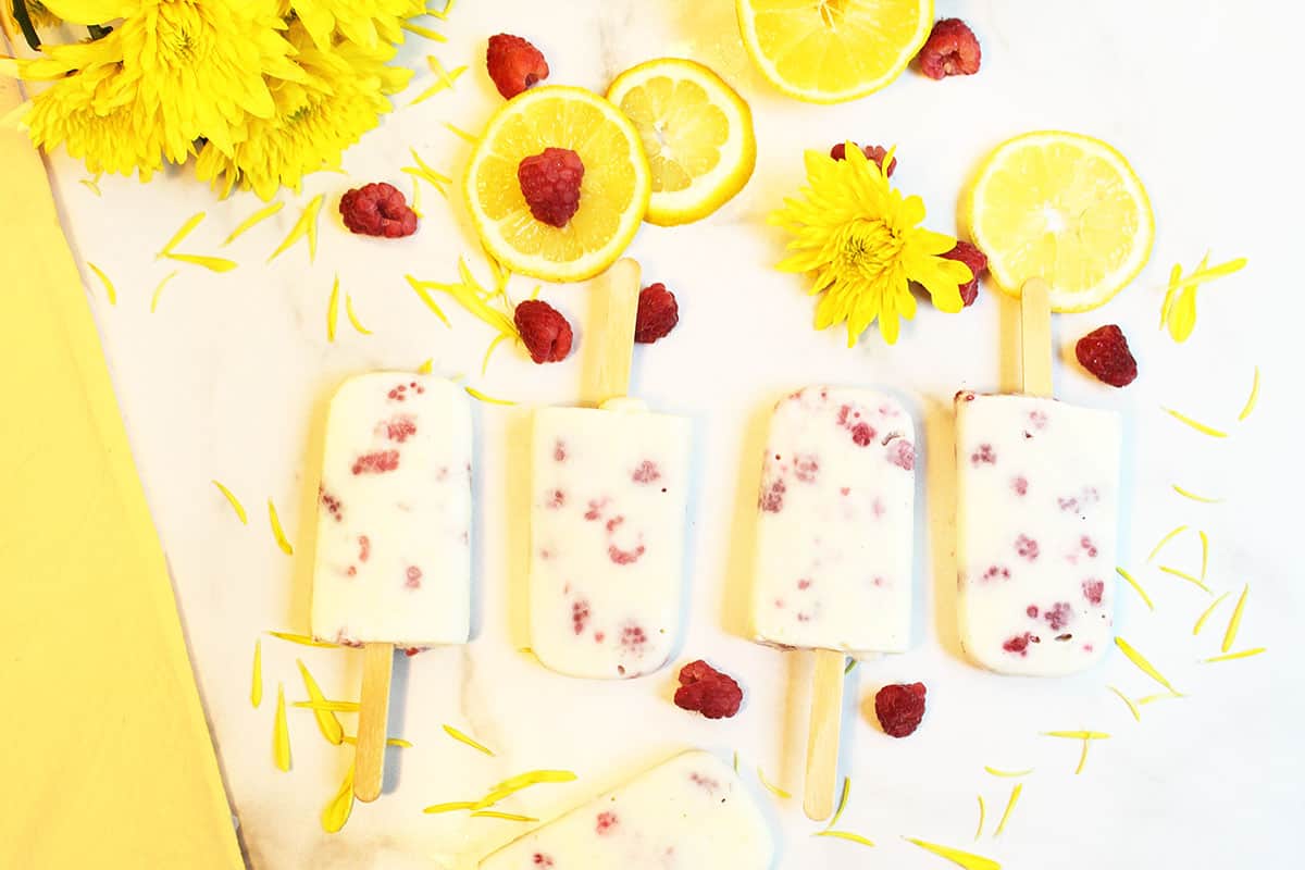 Popsicles lined up on white marble with yellowflowers napkin and raspberries around them.