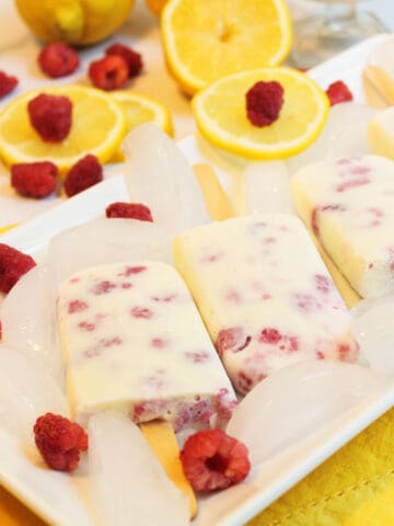 Popsicles on white platter with ice cubes and lemons and yogurt in serving cup behind it.