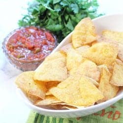 Closeup of tortilla chips in white bowl beside salsa and cilantro.