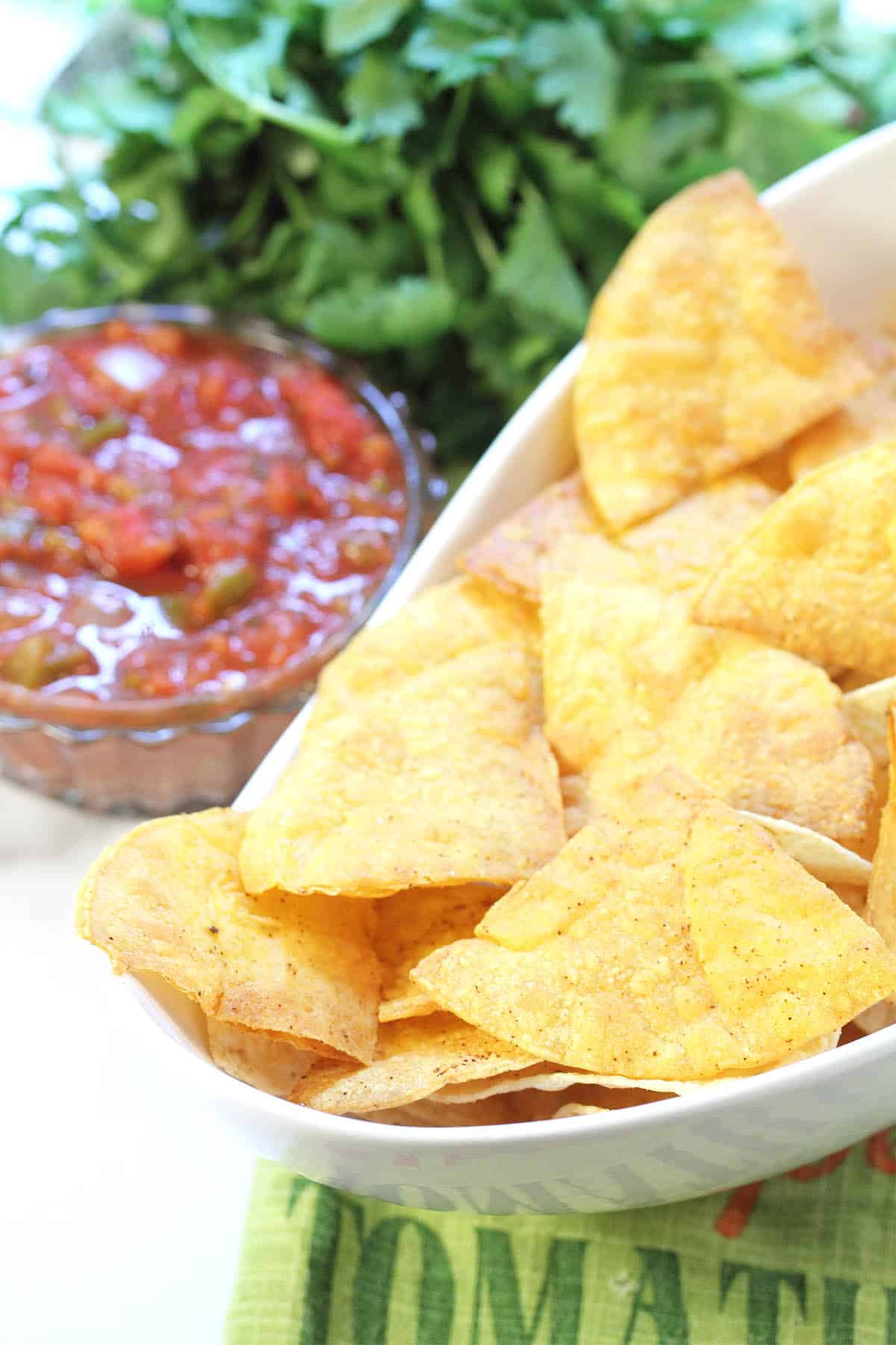 Closeup of tortilla chips in white bowl beside salsa and cilantro.