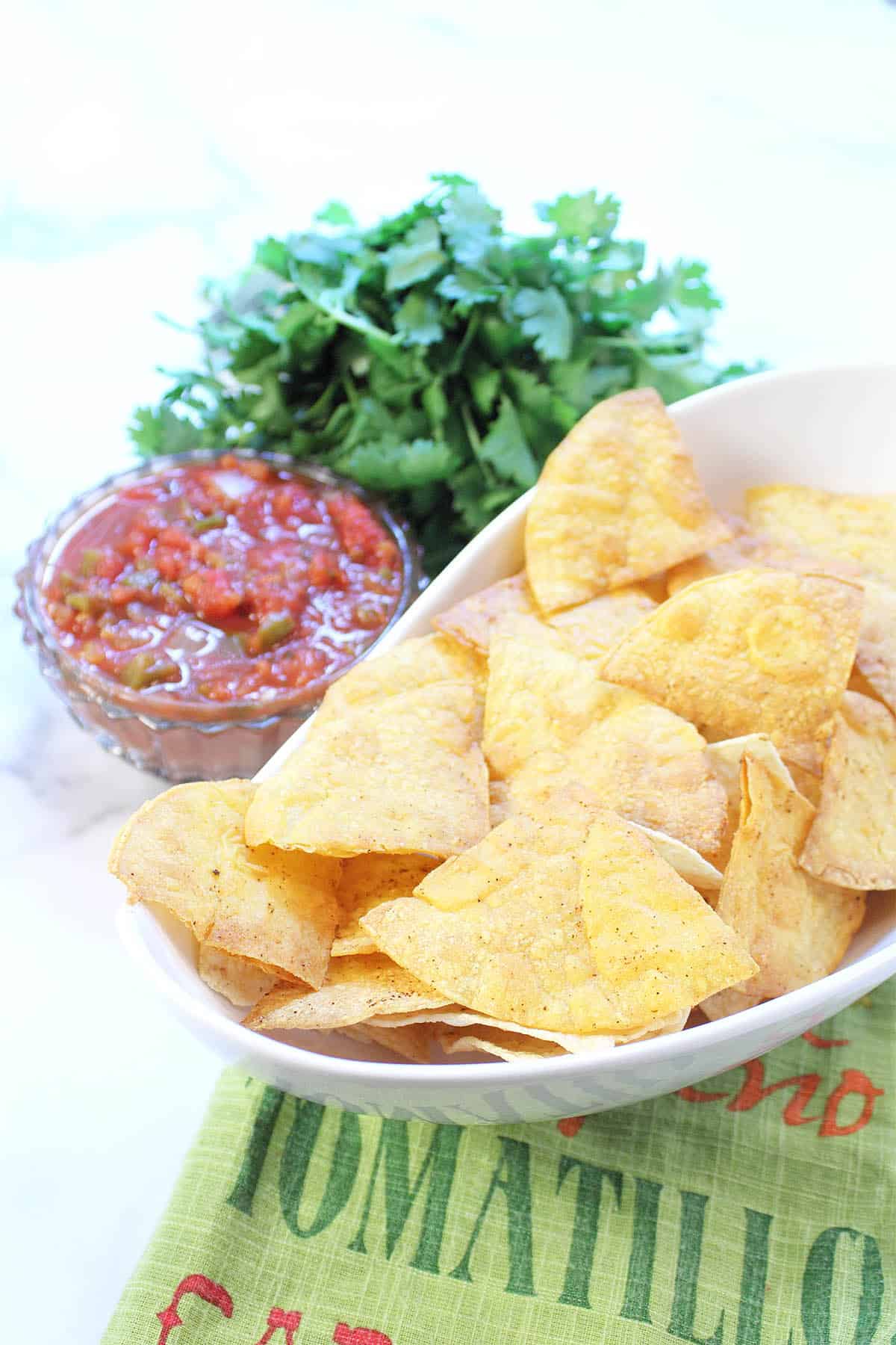Closeup of tortilla chips in white bowl beside salsa and cilantro.
