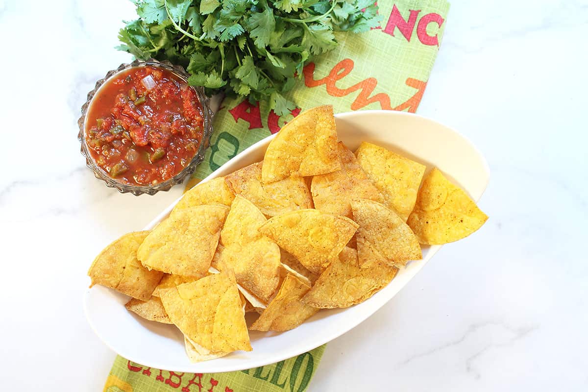 Overhead of curvy white bowl with baked chips on green towel with salsa on side.