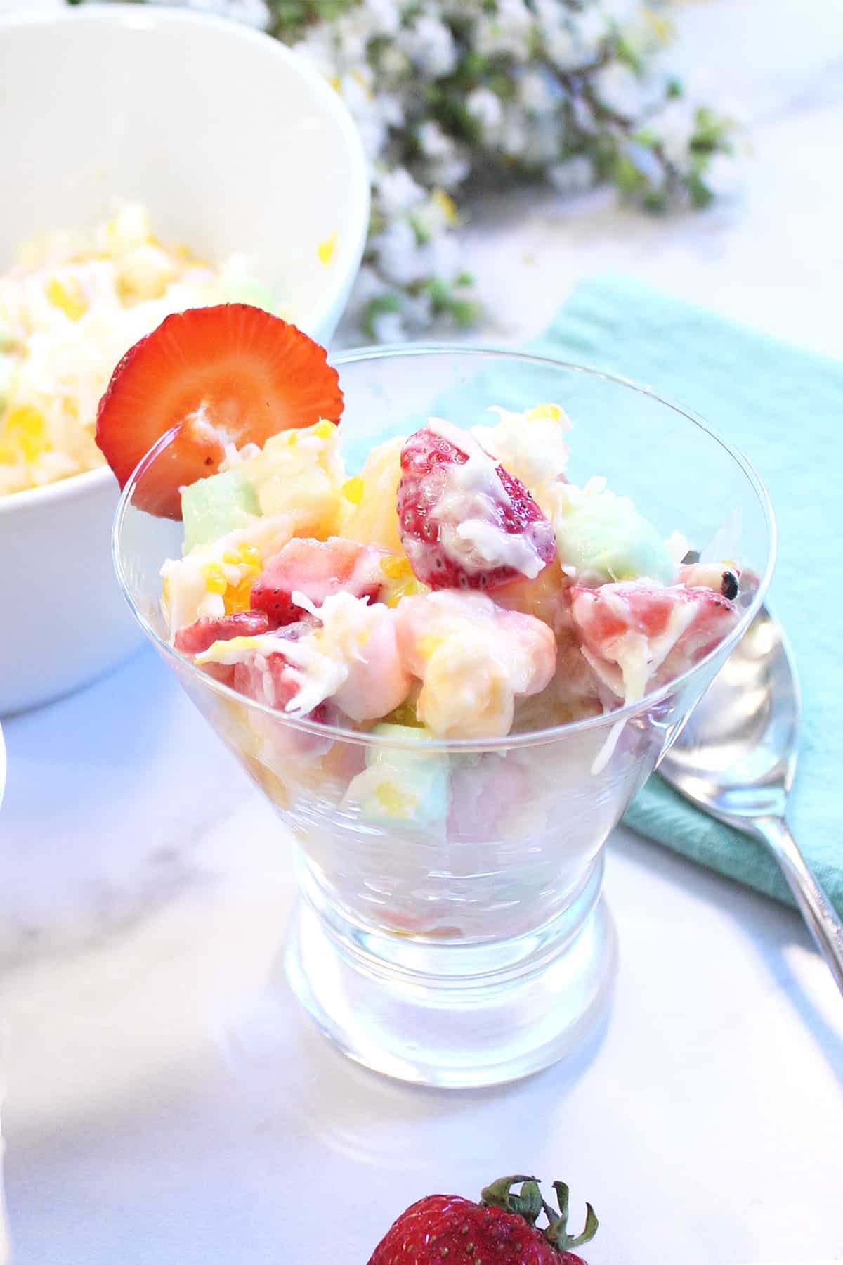 Single serving of Ambrosia Salad in glass dish on white table in front of serving bowl.