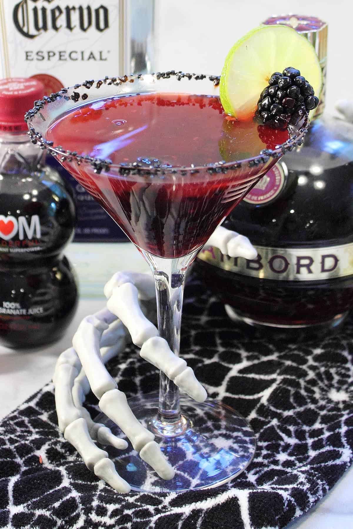 Closeup of skeleton hand grabbing blackberry margarita with ingredients around it.