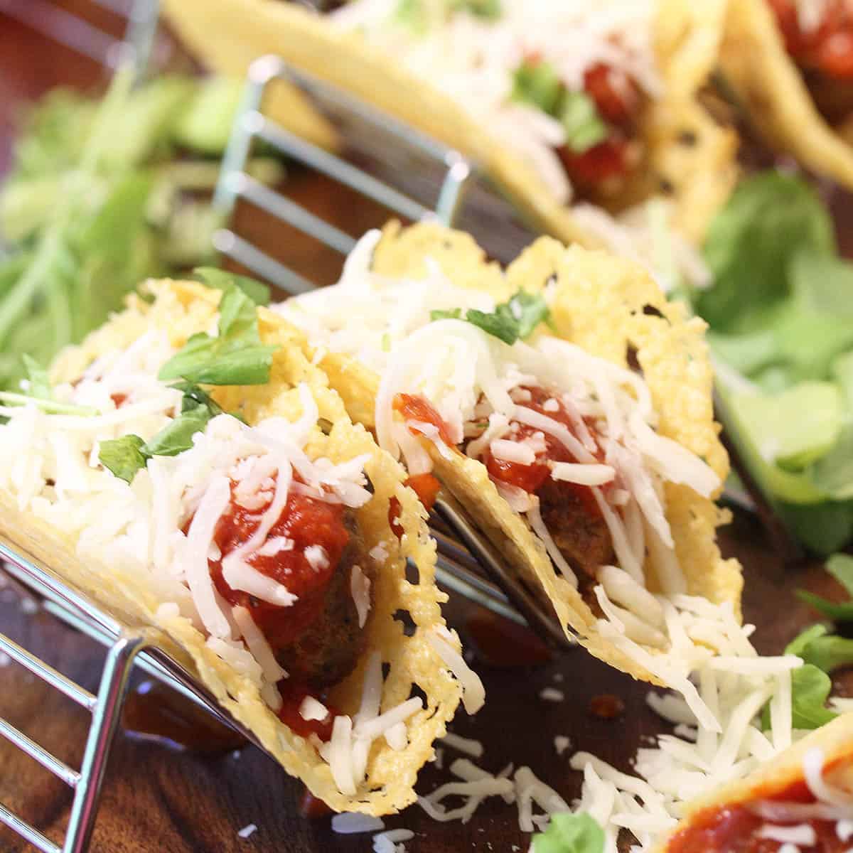 Closeup of 2 tacos with topping of cheese and garnish of chopped basil in taco stand.