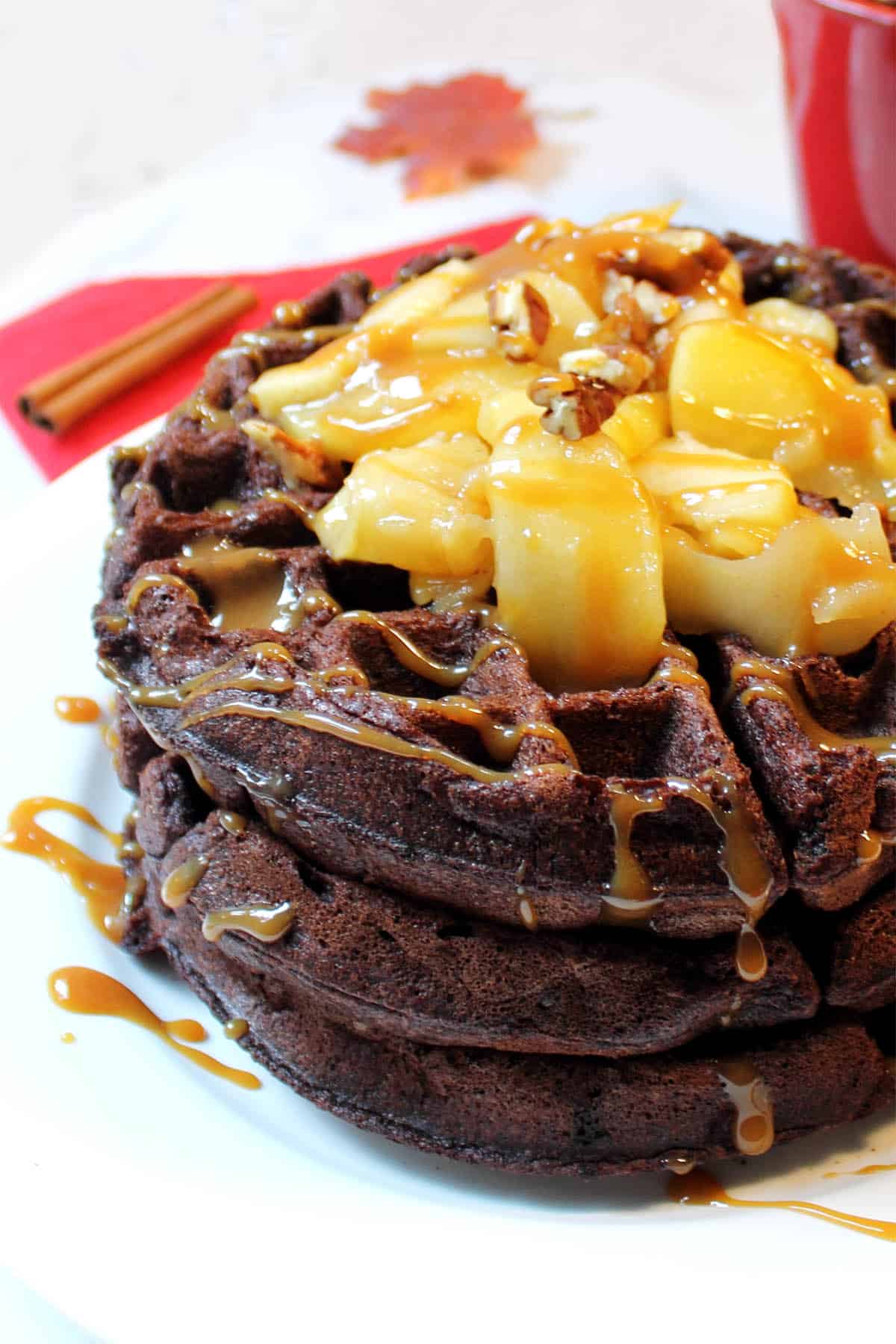 Partial view stack of chocolate Belgium waffles on white plate with sauteed apples and caramel sauce over top.