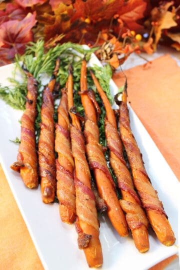 Bacon wrapped carrots on white platter with fall leaves in back.