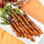 Bacon Wrapped Carrots on white platter with carrot greens on orange napkin.