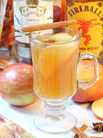 Warm apple cider cocktail with apples and liquor behind it.