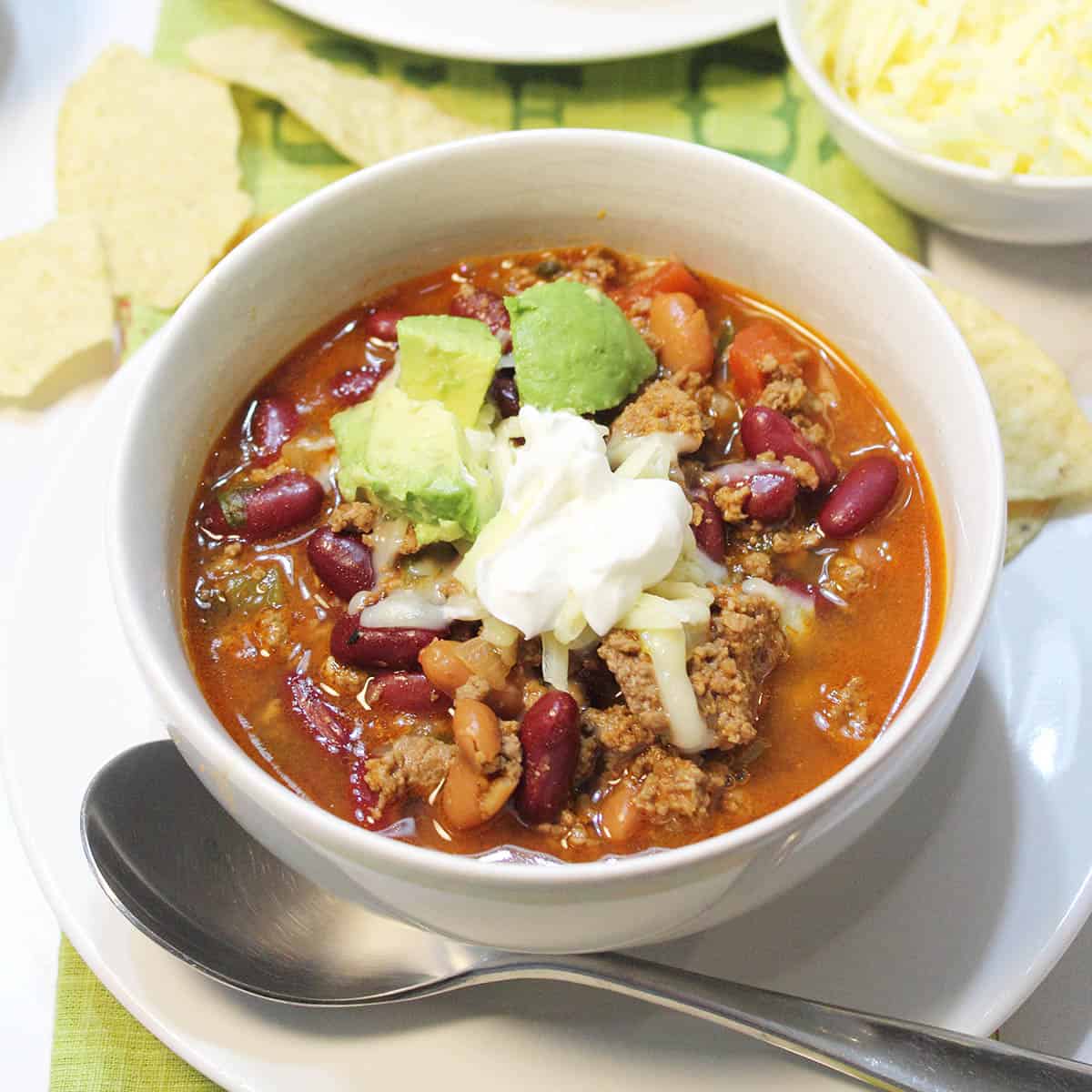 Bowl of chili on white table garnished with sour cream and avocado.