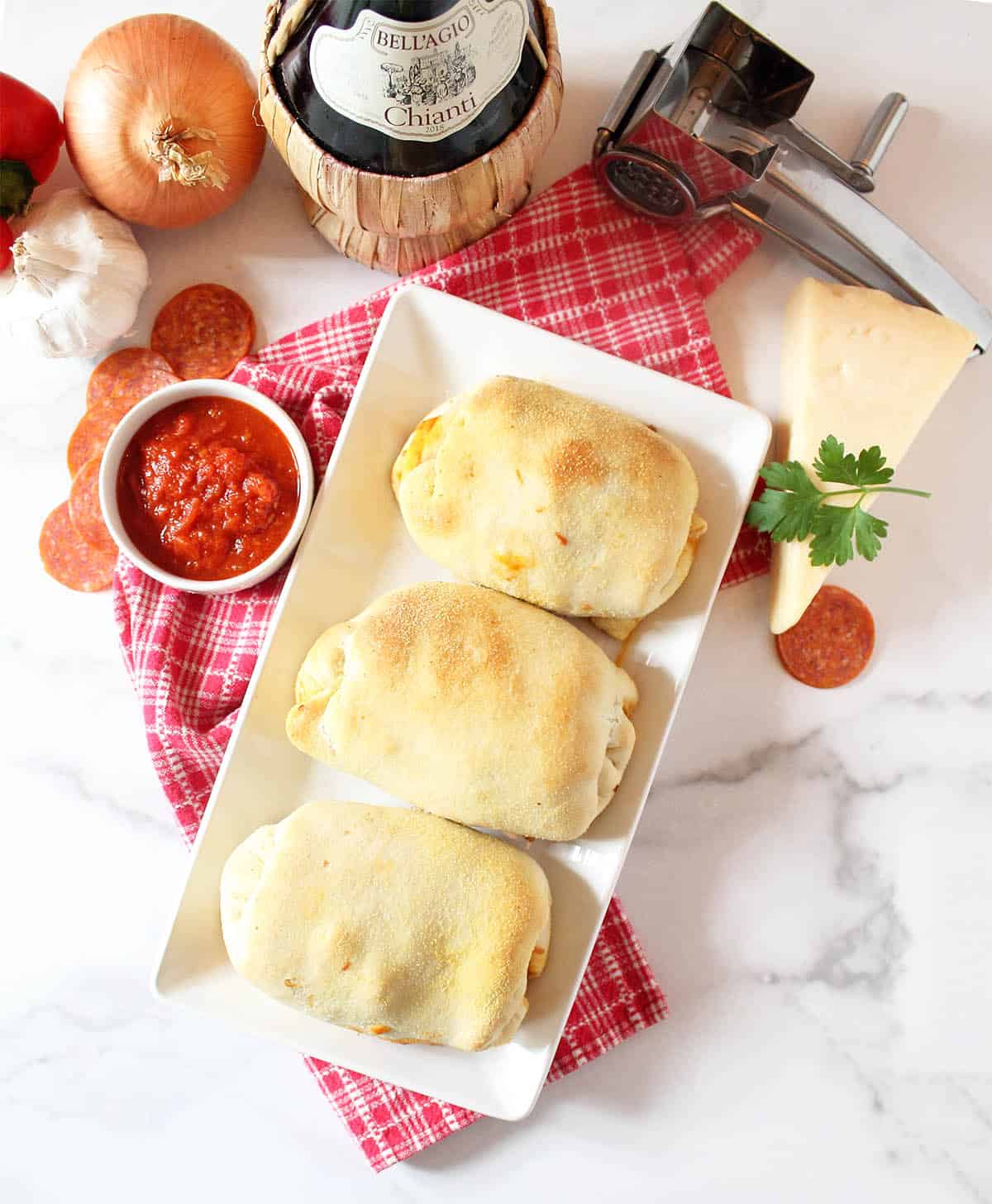 Overhead of baked stromboli on white platter with red checked towel and ingredients around them.