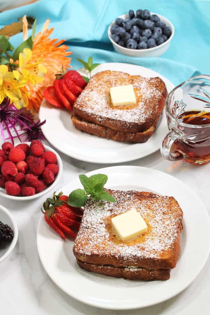 Two servings of Stuffed French Toast on white plates with berries, flowers and syrup beside them.