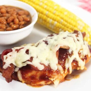 Closeup of bbq chicken breast on white plate with corn and baked beans.