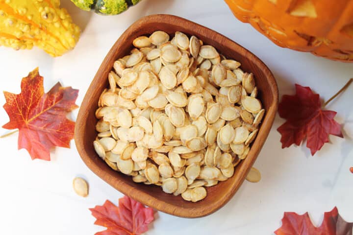 Overhead of pumpkin seeds in wooden bowl on white table with fall leaves.