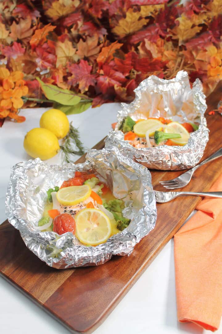 Two cooked salmon in foil packet dinners on wooden board.