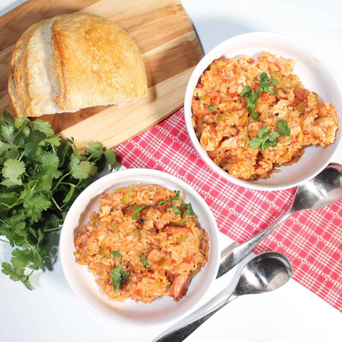 Overhead of 2 white bowls of sausage jambalaya with bread and red checkered towel.