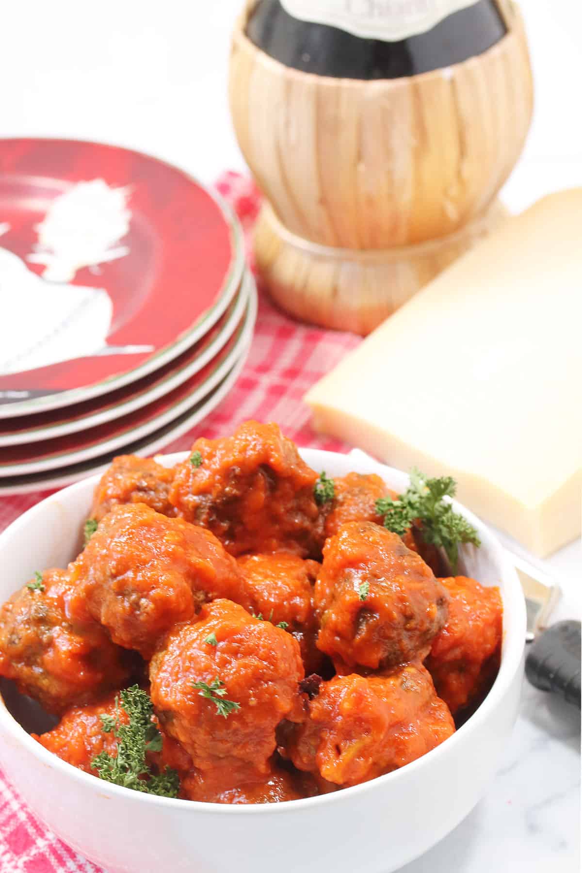 Meatballs covered in marinara in white bowl with cheese, wine bottle and serving plates.