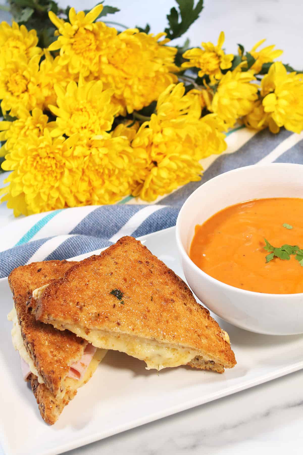 Sandwich next to bowl of tomato soup with yellow mums in back.