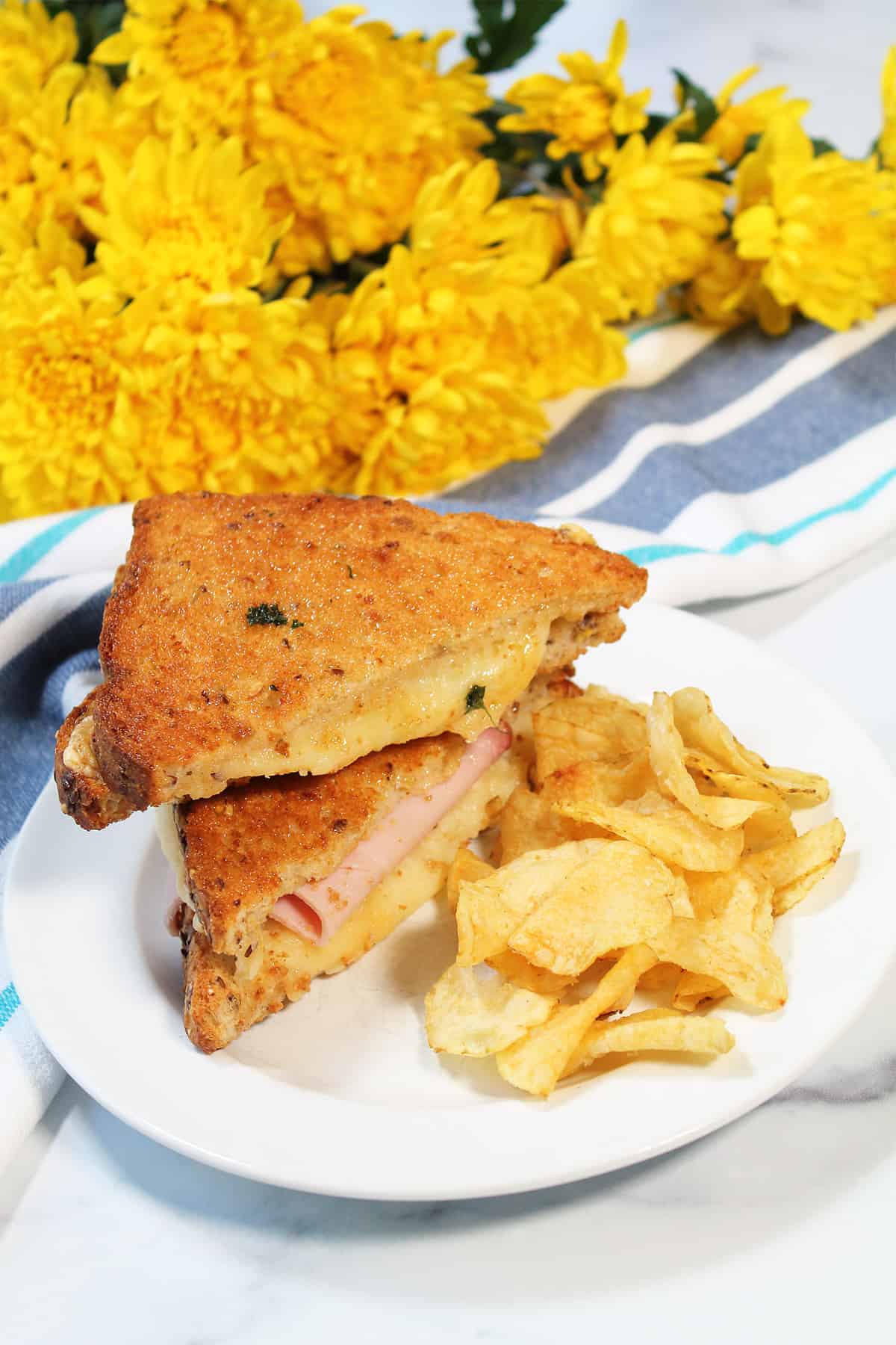 Grilled cheese sandwich with ham on white plate with potato chips.