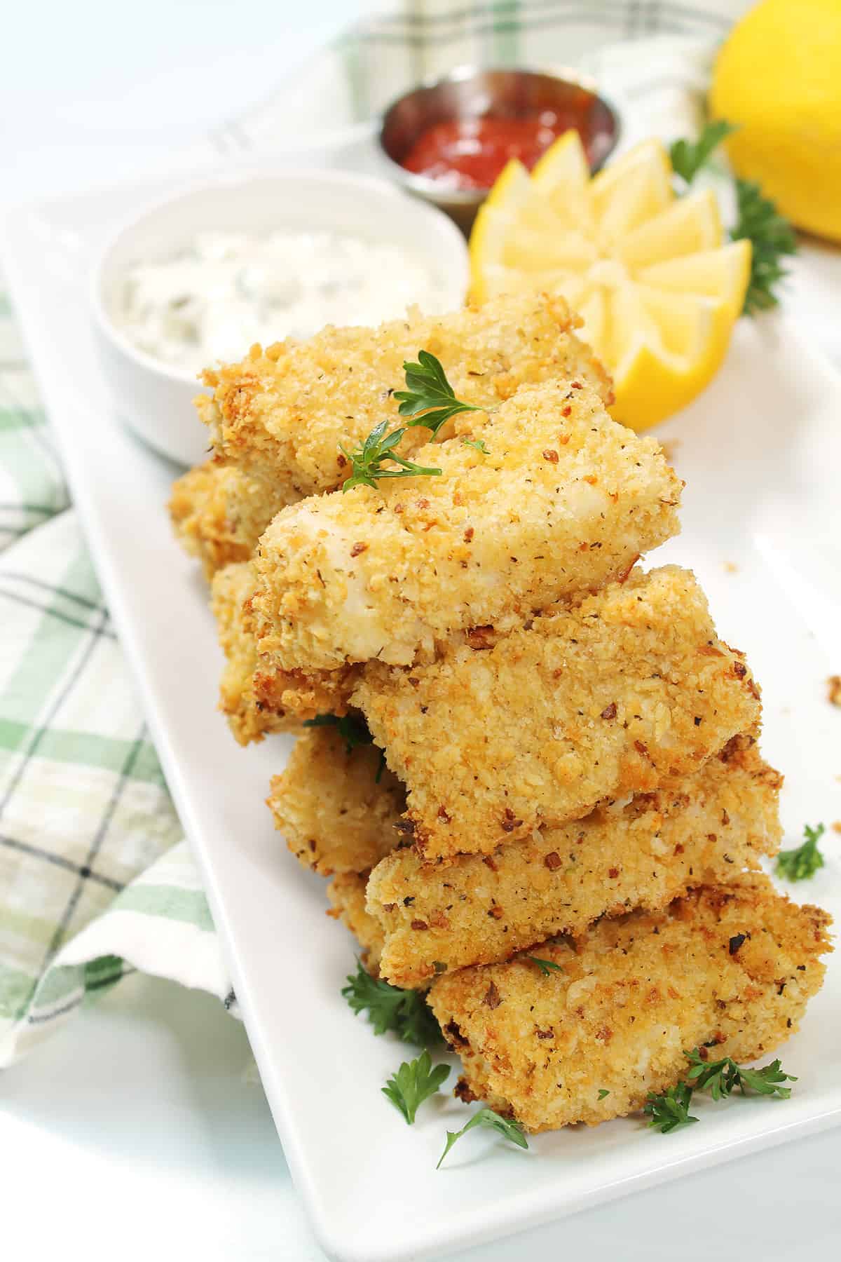 Stacked air fryer fish sticks on platter with tartar sauce and lemon wedge.